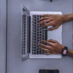 Top view of hands typing on a laptop beside a mask and smartphone.